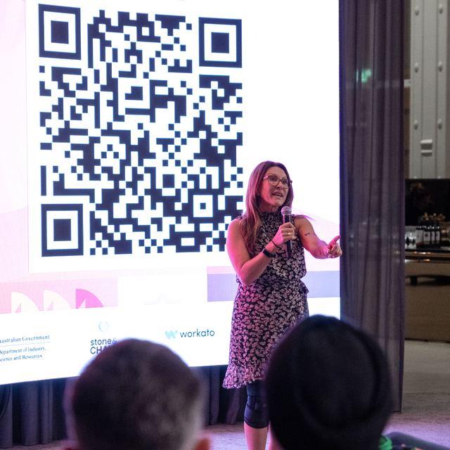 A speaker on stage beside a large QR code projected on the screen behind her