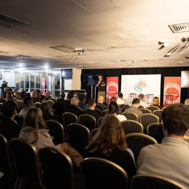 A full room of seated attendees facing a stage flanked by banners